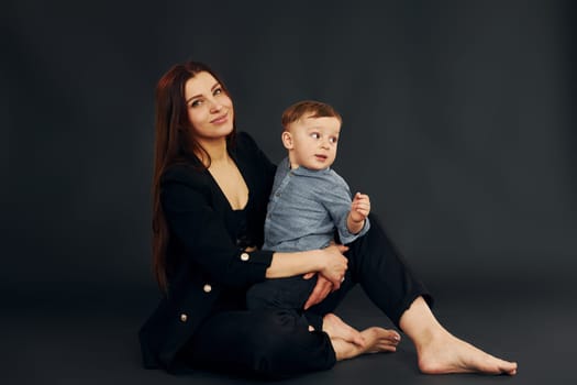 Sitting on the floor. Mother in stylish black clothes is with her little son in the studio.