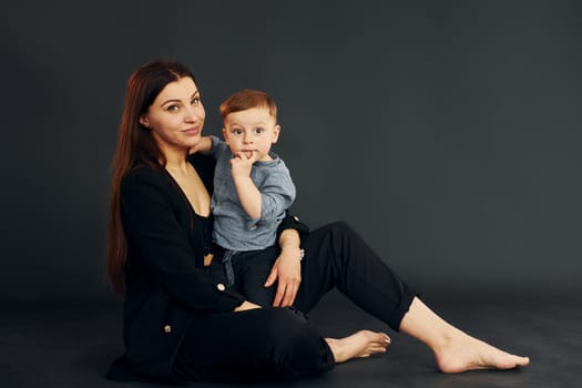 Sitting on the floor. Mother in stylish black clothes is with her little son in the studio.