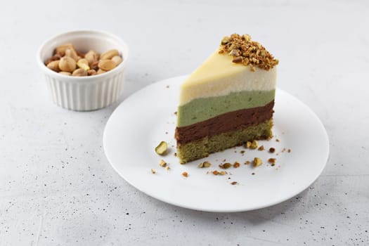 A piece of pistachio cake on a plate on a gray background. Ingredients: pistachio sponge cake, pistachio mousse, chocolate mousse and white chocolate mousse