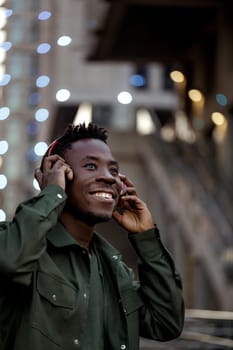 happy african-american man in stylish jacket in wireless headphones listening music on the street of the evening city