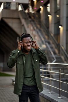 happy african-american man in stylish jacket in wireless headphones listening music on the street of the evening city
