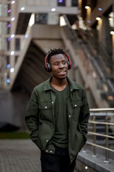 african-american man in stylish jacket in wireless headphones listening music on the street of the evening city