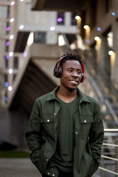 smiling african-american man in stylish jacket in wireless headphones listening music on the street of the evening city