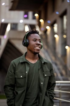 smiling african-american man in stylish jacket in wireless headphones listening music on the street of the evening city