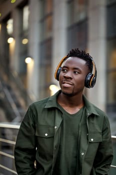 african-american man in stylish jacket in wireless headphones listening music on the street of the evening city