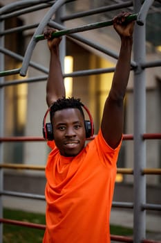 african-american man in orange t-hirt in wireless headphones pulling himself up on horizontal bars outdoor in the evening.