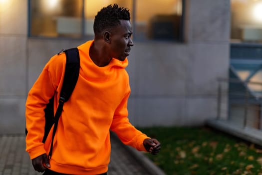 african-american man in stylish orange hoodie sweatshirt with backpack on city street in the evening