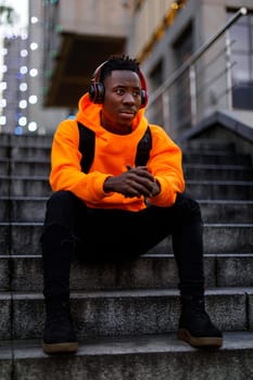 african-american man in stylish orange hoodie sweatshirt in wireless headphones listening music and sitting on stairs in city