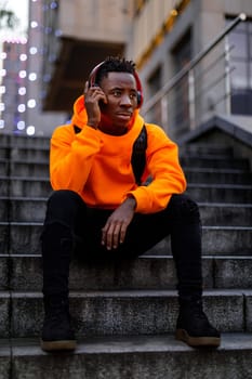 african-american man in stylish orange hoodie sweatshirt in wireless headphones listening music and sitting on stairs in city