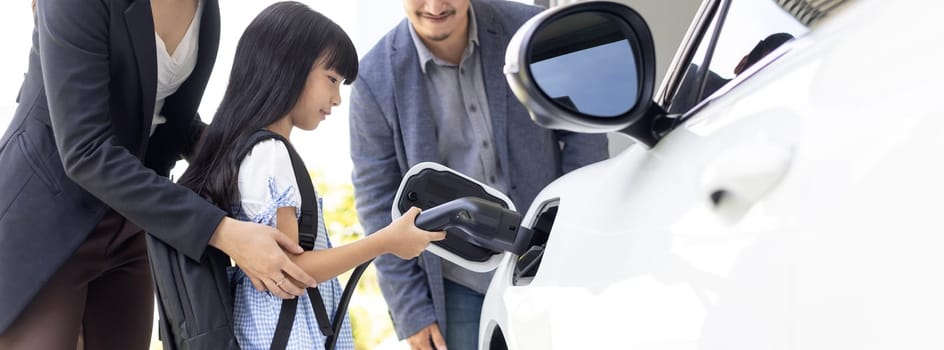 Progressive young parents and daughter with electric vehicle and home charging station. Green and clean energy from electric vehicles for healthy environment. Eco power from renewable source at home.