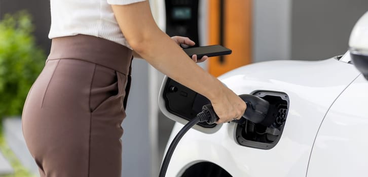 Closeup progressive woman install cable plug to her electric car with home charging station. Concept of the use of electric vehicles in a progressive lifestyle contributes to clean environment.