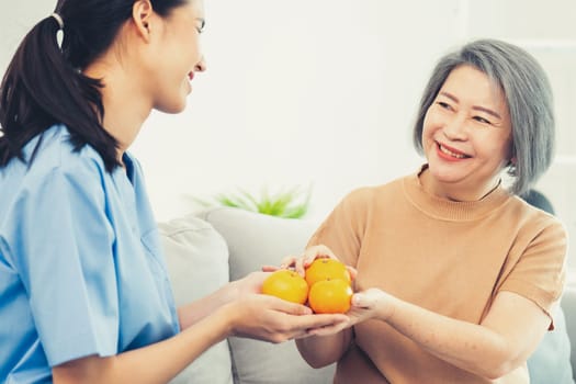 A young caregiver handing oranges to her contented senior patient at the living room. Senior care services, home visit by medical.