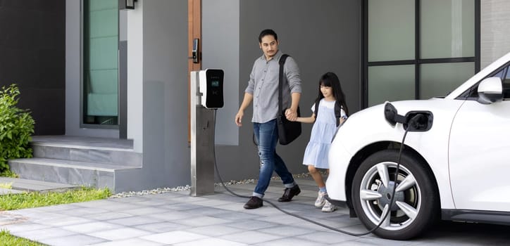 Progressive father and daughter plugs EV charger from home charging station to electric vehicle. Future eco-friendly car with EV cars powered by renewable source of clean energy.