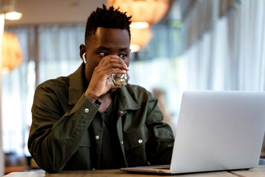 Cheerful young african american man in headphones using computer and drinking juice in cafe