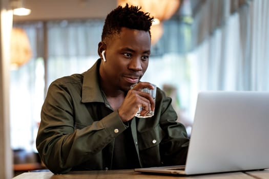 Cheerful young african american man in headphones using computer and drinking juice in cafe