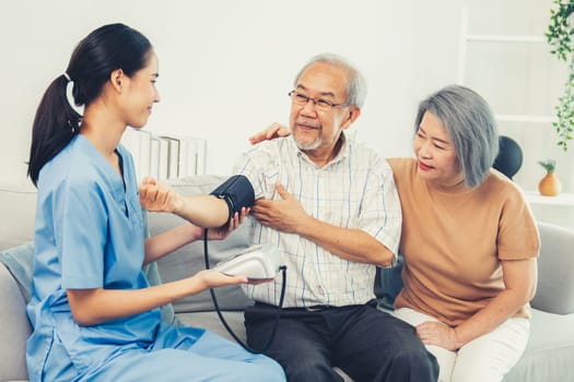 An elderly man having a blood pressure check by his personal caregiver with his wife sitting next to him in their home.