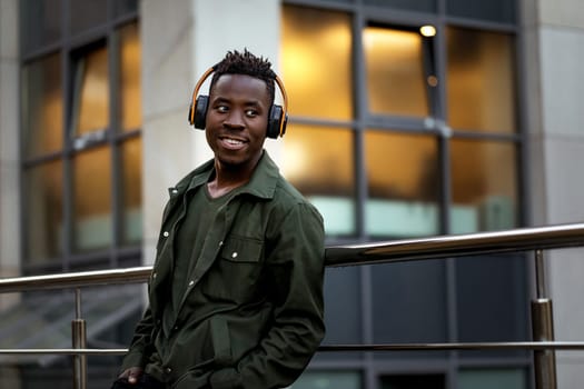 african-american man in stylish jacket in wireless headphones listening music on the street of the evening city. space for text