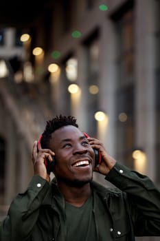 happy african-american man in stylish jacket in wireless headphones listening music on the street of the evening city