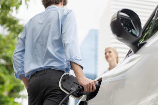 Focus hand insert EV charger to electric vehicle at public charging point in car park with blur business people in backdrop, eco-friendly lifestyle by rechargeable car for progressive concept.