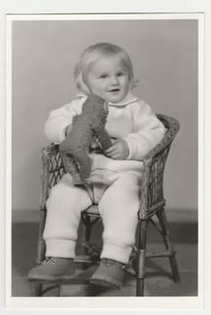 THE CZECHOSLOVAK SOCIALIST REPUBLIC - CIRCA 1970s: Vintage photo shows a small girl sits on wicker chair and holds plush toy - stuffed animal. Retro black and white photography. Circa 1970s.