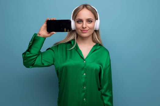 horizontal photo of stylish european woman holding smartphone with screen facing forward on blue studio background with free space.