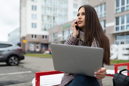 young caucasian woman manager talking on the phone holding a laptop on her knees sitting on a bench on the street.