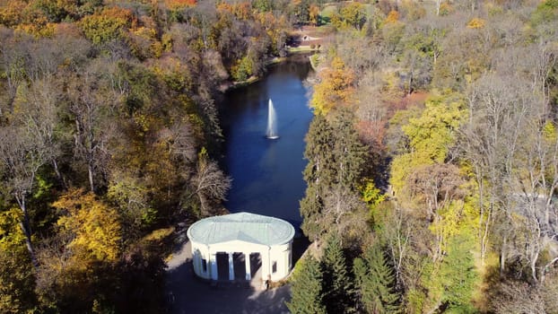 Beautiful landscaped park with lake, fountain, trees and white architecture on sunny autumn day. Trees with yellow green leaves on shores and high flowing jet of fountain. Aerial drone view