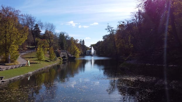 Beautiful landscaped park with decorative fountain in center of lake, trees and large stones on banks on sunny autumn day. Flying over water with fallen leaves and blue sky reflection. Many tourists