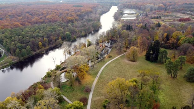 Panoramic view of large natural park with trees with yellow green red dry fallen leaves, river, forest, meadow and people walking dirt park paths on autumn cloudy day. Beautiful nature background.