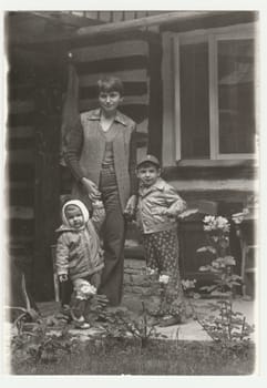 THE CZECHOSLOVAK SOCIALIST REPUBLIC - CIRCA 1980s: Vintage photo shows an adolescent girl with two children. They pose in front of log cabin. Retro black and white photography. Circa 1980s.