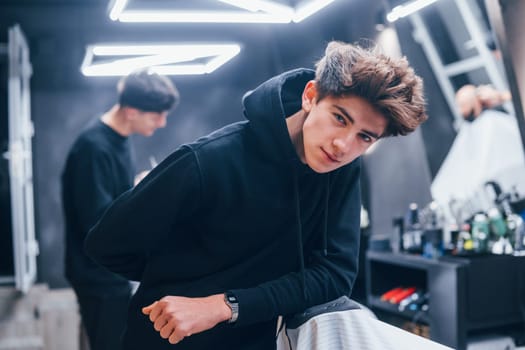 Young barber in black clothes standing in the barbershop. Workers and customer behind.