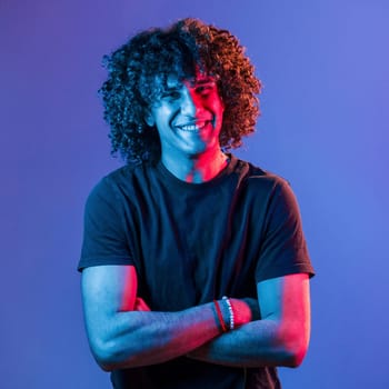 Posing for a camera. Young beautiful man with curly hair is indoors in the studio with neon lighting.