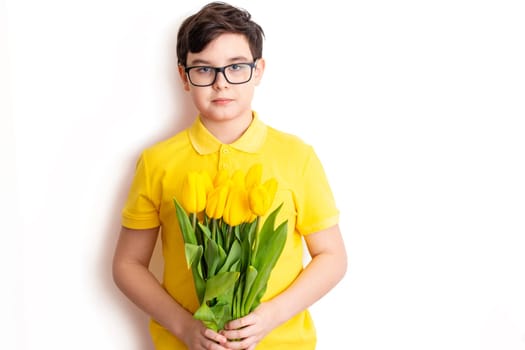 A cute boy in glasses and a yellow T-shirt holds a bouquet of yellow tulips, stands on a white background, isolated. Close up. Copy space.