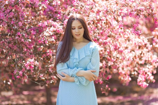 Alone beautiful girl, in light blue dress, standing in a pink blooming garden, looking away. Close up. Copy space