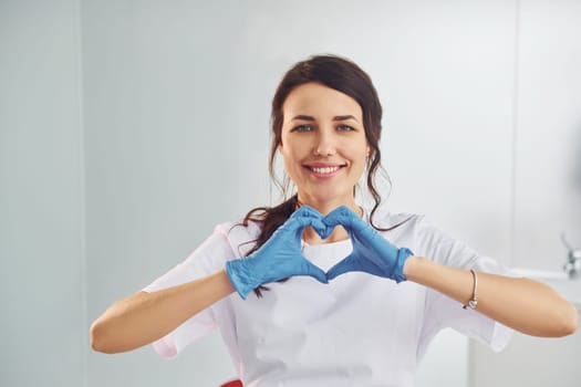 Heart shaped gesture. Portrait of professional female dentist with equipment that standing indoors.