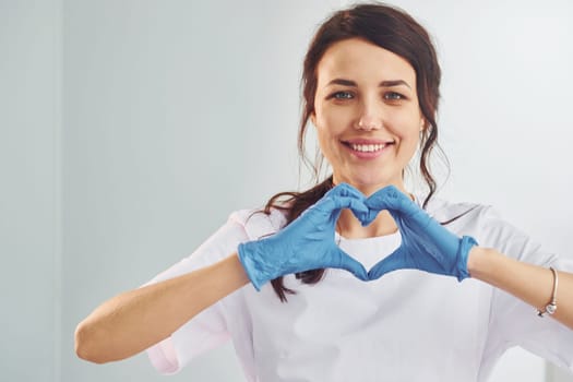 Heart shaped gesture. Portrait of professional female dentist with equipment that standing indoors.