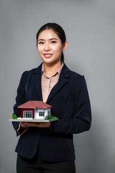 Young Asian woman wearing formal suit, holding and showcasing house model on isolated background. Ideal for housing advertisement, home insurance promotions, or customizable copy space. Enthusiastic
