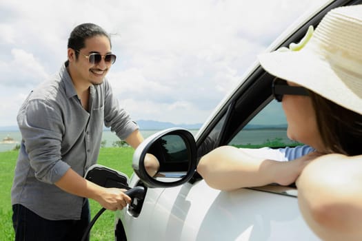 Concept of progressive happy young couple relax and enjoy natural green field and lake with electric vehicle. Electric vehicle driven by clean renewable. Refreshing environment from green energy.