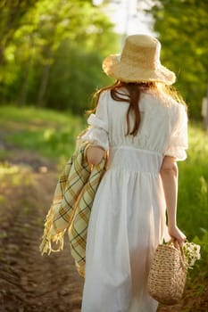 a woman in a light dress and a wicker hat with a basket and a plaid in her hands walks through the forest in sunny weather. Photo from the back. High quality photo