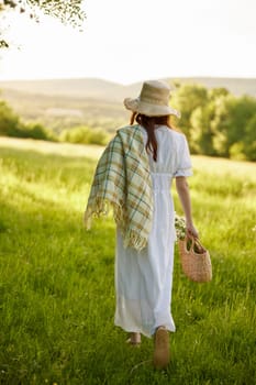 a woman in a light dress and a wicker hat with a basket and a plaid in her hands walks through the forest in sunny weather. Photo from the back. High quality photo