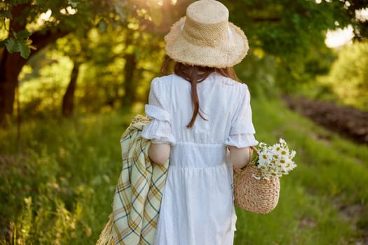 a woman in a light dress and a wicker hat with a basket and a plaid in her hands walks through the forest in sunny weather. Photo from the back. High quality photo