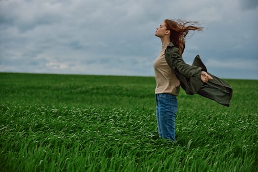 a red-haired woman in a long coat stands in a green field and the wind blows her hair. High quality photo