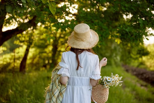 a woman in a light dress and a wicker hat with a basket and a plaid in her hands walks through the forest in sunny weather. Photo from the back. High quality photo