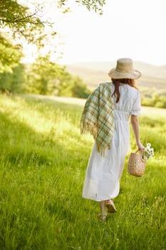 a woman in a light dress and a wicker hat with a basket and a plaid in her hands walks through the forest in sunny weather. Photo from the back. High quality photo