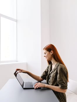 a slender, red-haired woman works at a laptop in a bright office room. High quality photo