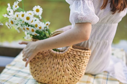 a woman sits with her back to the camera by the lake holding a wicker basket with daisies in her hand. subject photography without a face. High quality photo