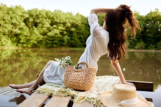 an elegant woman in a light dress sits on a pier by the lake with her legs in the water and looking at the landscape straightens her hair turning away from the camera. High quality photo