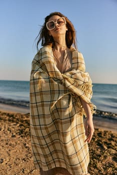 a cute woman stands with a plaid during sunset on the beach and the wind blows her red hair. High quality photo