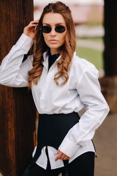 A stylish girl in a white shirt and sunglasses standing on the street during the day.