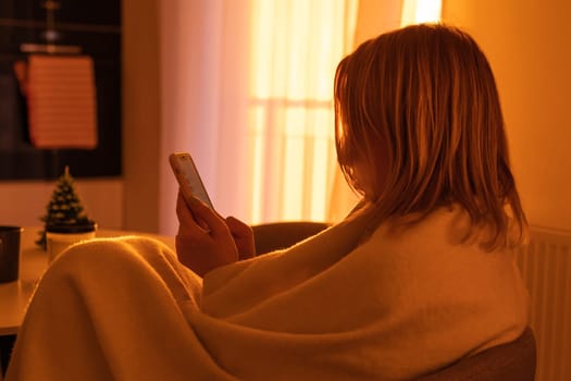 A girl wrapped in a blanket uses a mobile phone in a romantic atmosphere in an apartment with a cozy sunset light from the window, side view